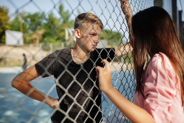 Porträt eines jungen coolen Jungen, der auf einem Basketballplatz steht und nachdenklich ein hübsches Mädchen mit dunklen Haaren durch einen Maschendrahtzaun betrachtet