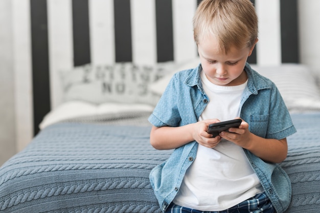 Porträt eines Jungen, der vor Bett unter Verwendung des Handys steht