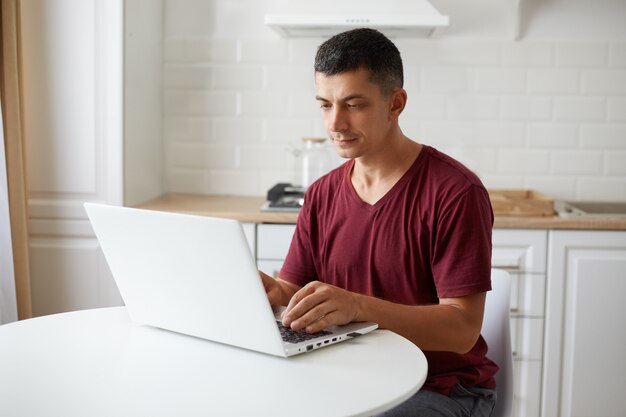 Porträt eines jungen erwachsenen mannes, der ein kastanienbraunes t-shirt im casual-stil trägt, freiberufler, der von zu hause aus online arbeitet, am tisch in der küche sitzt und konzentriert auf die notebook-anzeige schaut.