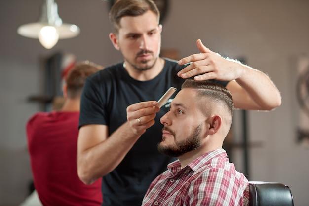Porträt eines jungen Friseurs, der Haare seines bärtigen Kunden stylt.