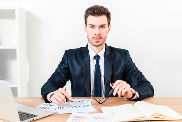 Porträt eines jungen Geschäftsmannbehälters über dem Diagramm und den Brillen in der Hand, die zur Kamera schauen