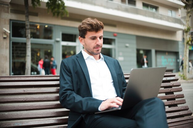 Porträt eines jungen Geschäftsmannes, der auf Bank unter Verwendung des Laptops sitzt