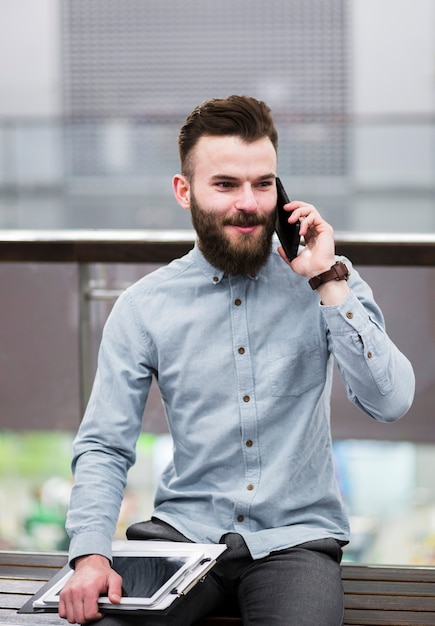 Porträt eines jungen Geschäftsmannes, der auf der Bank hält das Klemmbrett und digitale Tablette spricht am Handy sitzt