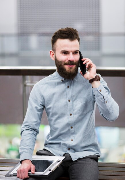 Porträt eines jungen Geschäftsmannes, der auf der Bank hält das Klemmbrett und digitale Tablette spricht am Handy sitzt