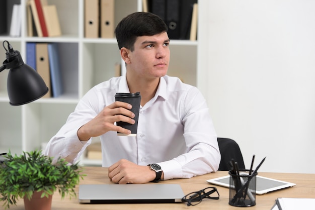 Porträt eines jungen Geschäftsmannes, der in der Hand Wegwerfkaffeetasse am Schreibtisch hält