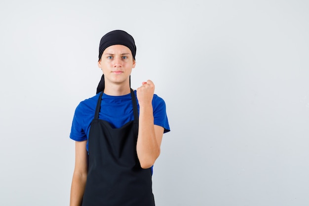 Porträt eines jungen Kochmannes, der die geballte Faust in T-Shirt, Schürze hebt und ernsthafte Vorderansicht sieht