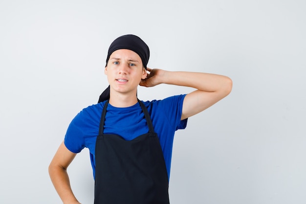 Porträt eines jungen Kochmannes, der sich am Kopf kratzt, während er die Hand in T-Shirt, Schürze und nachdenklicher Vorderansicht auf der Hüfte hält