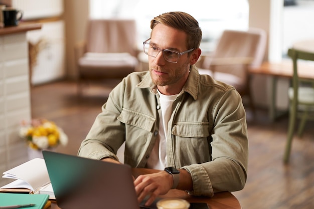 Kostenloses Foto porträt eines jungen konzentrierten mannes in brille, der von einem café aus arbeitet und nachdenklich studiert