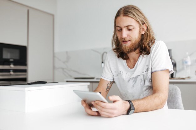 Porträt eines jungen lächelnden Mannes, der zu Hause in der Küche sitzt und ein digitales Tablet verwendet