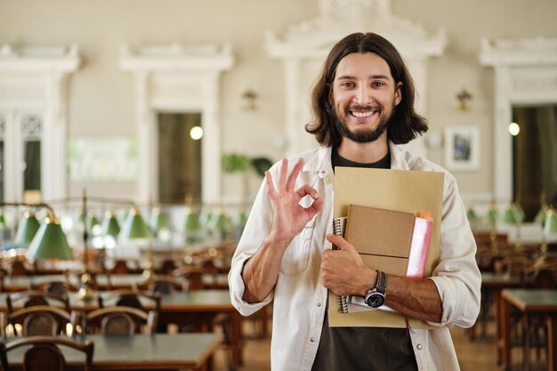 Porträt eines jungen, lächelnden, zufälligen männlichen Studenten mit einem Buch, das in der Bibliothek der Universität glücklich eine OK-Geste zeigt