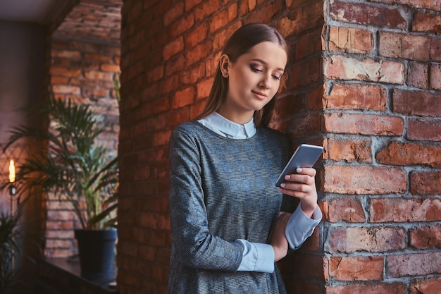 Porträt eines jungen Mädchens in einem eleganten grauen Kleid, das sich mit einem Smartphone an die Ziegelwand lehnt.