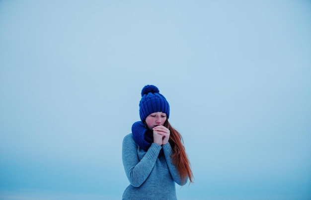 Porträt eines jungen Mädchens mit roten Haaren und Sommersprossen, das im Hintergrundeis des Wintertages eine blau gestrickte Wollmütze und einen Schal trägt