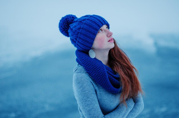 Porträt eines jungen Mädchens mit roten Haaren und Sommersprossen, das im Hintergrundeis des Wintertages eine blau gestrickte Wollmütze und einen Schal trägt