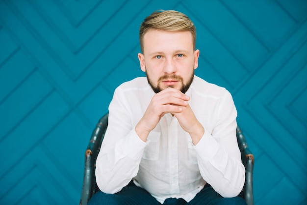 Kostenloses Foto porträt eines jungen mannes, der auf stuhl mit der hand sitzt, umklammerte gegen blaue designwand