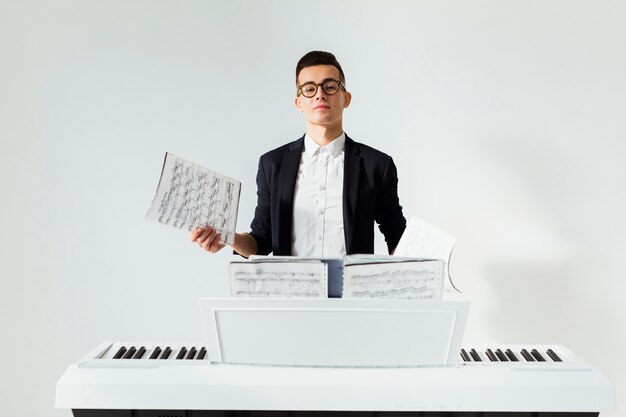 Porträt eines jungen Mannes, der das musikalische Blatt steht hinter dem Klavier gegen weißen Hintergrund hält