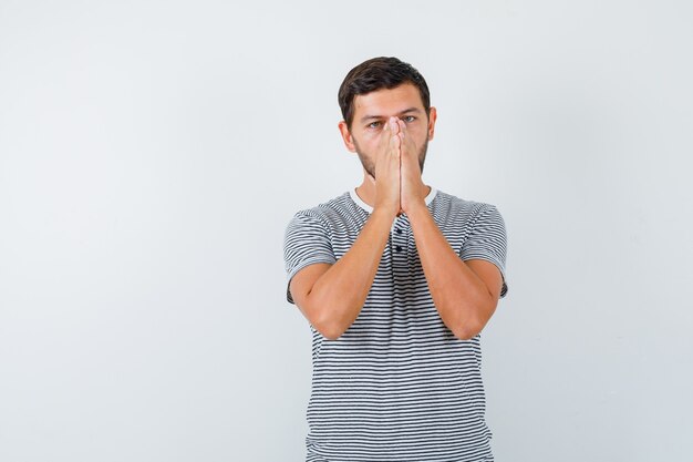 Porträt eines jungen Mannes, der die Hände zusammendrückt, während er im T-Shirt betet und hoffnungsvolle Vorderansicht schaut