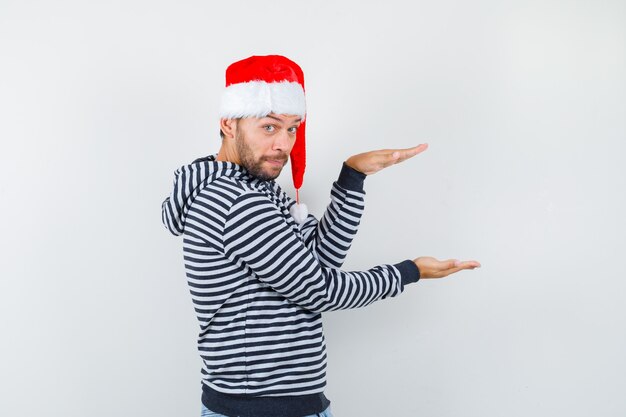 Porträt eines jungen Mannes, der ein Größenschild in Hoodie, Weihnachtsmütze und zögerlichem Blick zeigt