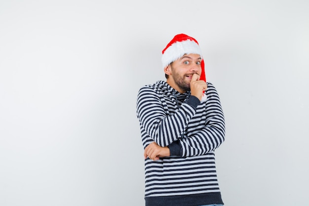 Porträt eines jungen mannes, der hand am kinn in hoodie, weihnachtsmütze hält und interessiert aussieht