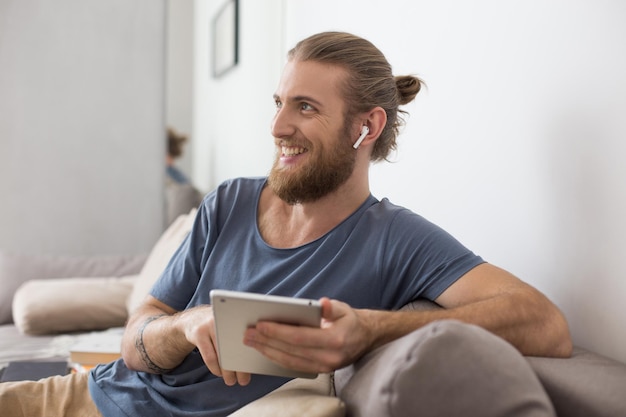 Porträt eines jungen Mannes, der mit Kopfhörern und dem Tablet in den Händen auf einem grauen Sofa sitzt und glücklich zu Hause beiseite schaut