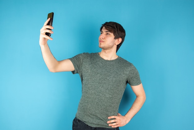 Porträt eines jungen Mannes, der selfie mit Handy gegen Blau nimmt.