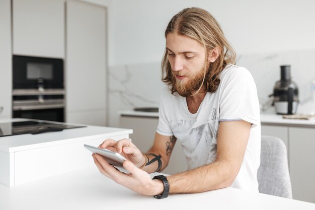 Porträt eines jungen Mannes, der zu Hause in der Küche sitzt und ein digitales Tablet verwendet