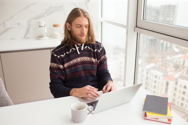 Porträt eines jungen Mannes im Pullover, der mit einer Tasse am Tisch sitzt und zu Hause am Laptop arbeitet