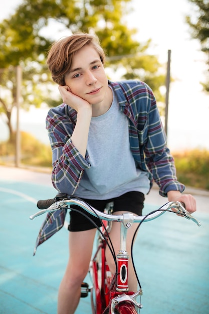Porträt eines Jungen mit blonden Haaren in Shorts und Freizeithemd, der mit rotem Fahrrad auf dem Basketballplatz im Park steht Junger Mann, der verträumt in die Kamera blickt, während er mit einem klassischen Fahrrad steht