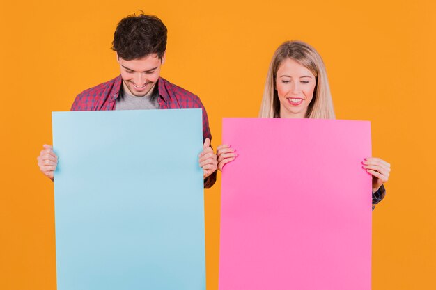 Porträt eines jungen Paares, das blaues und rosa Plakat gegen einen orange Hintergrund betrachtet