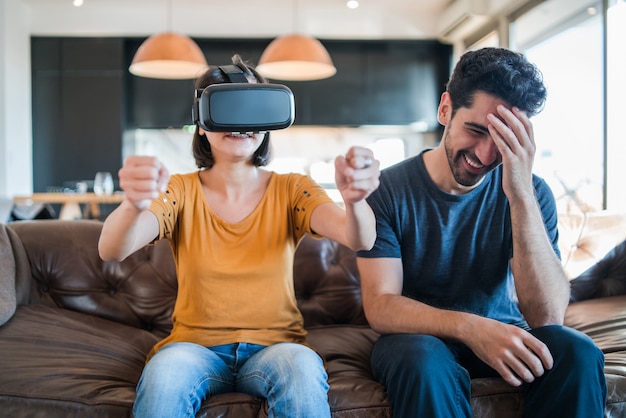 Porträt eines jungen Paares, das zusammen Spaß hat und Videospiele mit VR-Brille spielt, während es zu Hause auf der Couch sitzt