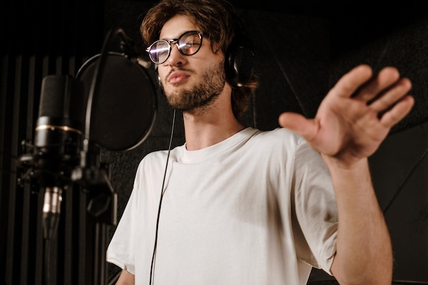 Porträt eines jungen Sängers mit Kopfhörern, der während der Aufnahme eines neuen Songs im modernen Studio singt