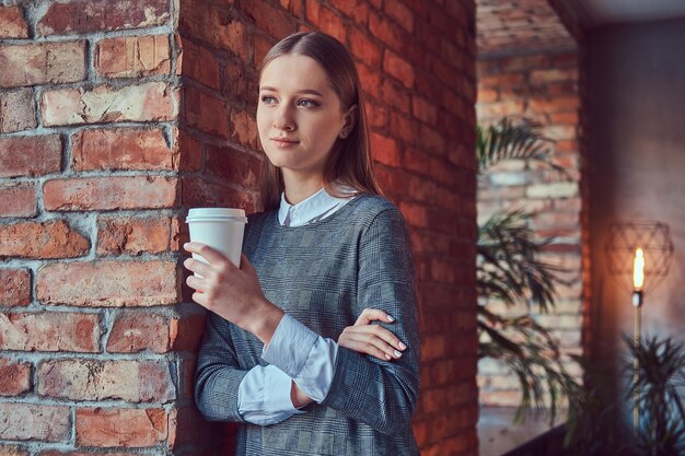 Porträt eines jungen, schlanken, sinnlichen Mädchens in einem grauen Kleid, das sich mit einem Morgenkaffee an eine Ziegelwand lehnt.