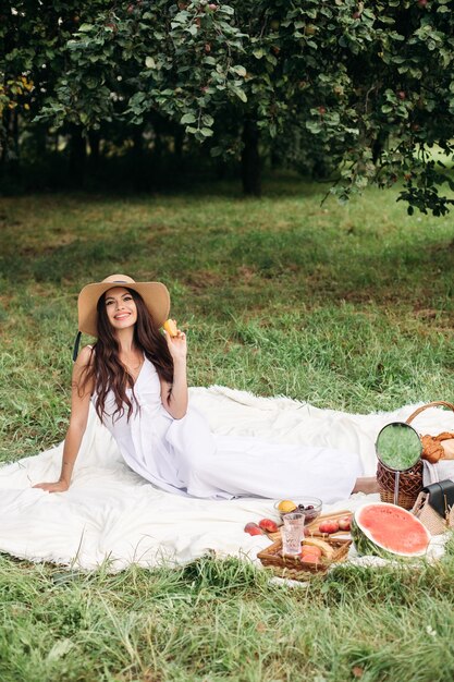Porträt eines jungen schönen Mädchens mit gleichmäßigen weißen Zähnen, einem schönen Lächeln in einem Strohhut und einem langen weißen Kleid machen ein Picknick im Garten