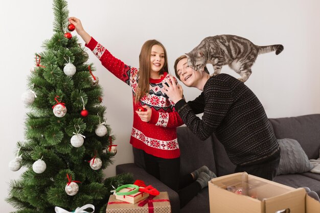 Porträt eines jungen schönen Paares mit einer Katze, die zu Hause steht und gemeinsam den Weihnachtsbaum schmückt