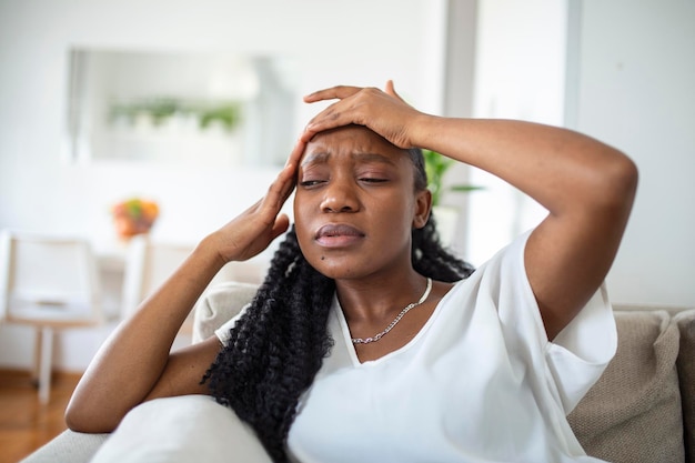 Porträt eines jungen schwarzen Mädchens, das zu Hause mit Kopfschmerzen und Schmerzen auf der Couch sitzt Schöne Frau, die unter chronischen täglichen Kopfschmerzen leidet Traurige Frau, die ihren Kopf hält, weil Nebenhöhlenschmerzen