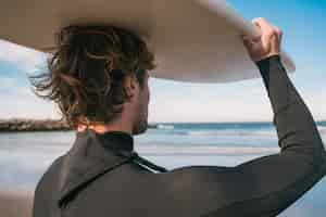 Kostenloses Foto porträt eines jungen surfers am strand, der sein surfbrett hochhält und einen schwarzen surfanzug trägt. sport- und wassersportkonzept.