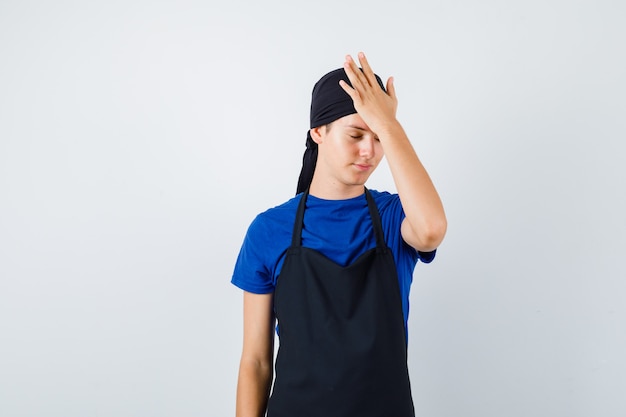 Porträt eines jungen Teenager-Kochs mit Hand auf dem Kopf in T-Shirt, Schürze und vergesslicher Vorderansicht