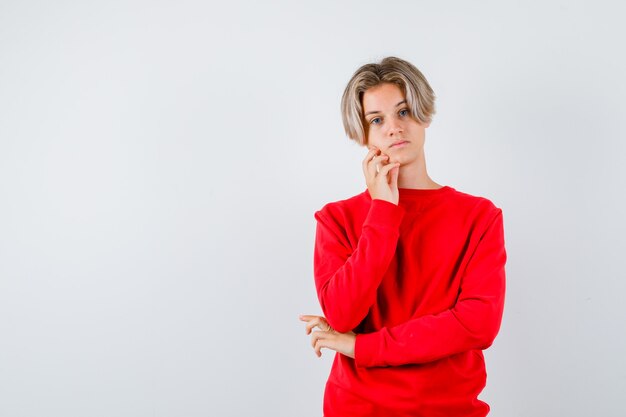 Porträt eines jungen Teenagers, der im roten Pullover die Finger auf die Wange hält und verärgerte Vorderansicht schaut