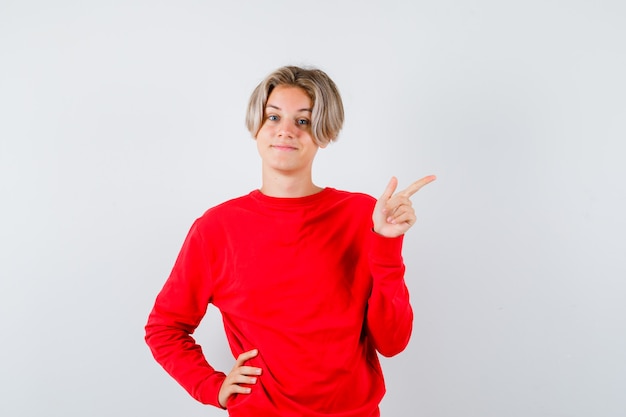 Porträt eines jungen Teenagers, der in rotem Pullover auf die obere rechte Ecke zeigt und fröhliche Vorderansicht sieht
