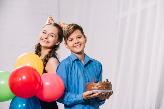 Porträt eines Jungen und des Mädchens, die bunte Ballone und Kuchen auf Platte halten