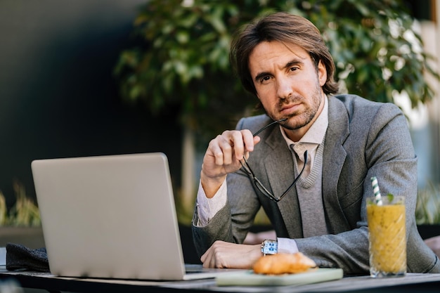 Porträt eines jungen Unternehmers, der in einem Café sitzt und am Laptop arbeitet, während er in die Kamera blickt.