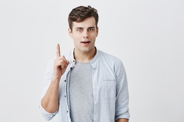 Porträt eines kaukasischen Mannes mit Käferaugen und dunklem Haar, der von etwas geschockt wird, gekleidet in ein blaues Hemd über einem T-Shirt, das seinen Finger auf eine leere Wand mit Kopierraum für Ihren Werbeinhalt zeigt