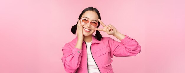 Porträt eines kawaii asiatischen Mädchens mit Sonnenbrille, das ein Friedenszeichen in der Nähe des Auges zeigt und süß in die Kamera p lächelt