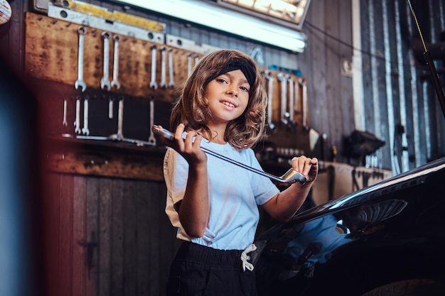 Porträt eines kleinen fröhlichen Mädchens mit großem Schraubenschlüssel in den Händen in der Nähe von glänzendem Gar beim Autoservice.