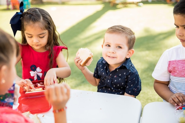 Kostenloses Foto porträt eines kleinen jungen, der während seiner mittagspause im kindergarten ein sandwich isst und lächelt