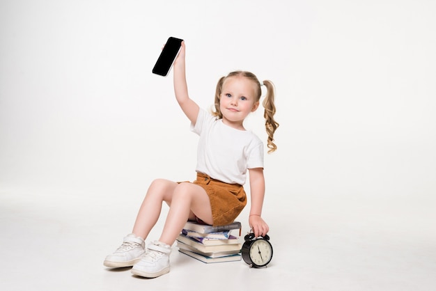 Porträt eines kleinen Mädchens, das Telefonbildschirm hält, der auf einem Stapel Bücher sitzt.