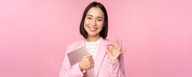 Porträt eines korporativen Frauenmädchens im Büro im Business-Anzug, das ein digitales Tablet hält, das in Ordnung ist und die Firma empfiehlt, die über rosa Hintergrund steht