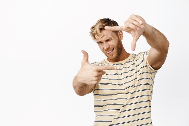 Porträt eines kreativen blonden Kerls, der sich vorstellt, wie er einen guten Schuss macht und nach dem perfekten Winkel mit der Geste des Handrahmens sucht, die etwas auf dem weißen Hintergrund des Auges misst