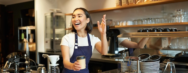 Kostenloses Foto porträt eines lächelnden asiatischen barista-mädchens, das im café bestellung aufgibt und gäste einlädt, essen zum mitnehmen abzuholen