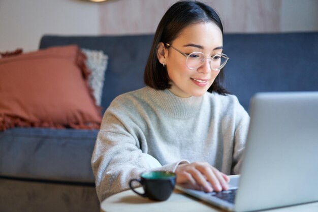 Porträt eines lächelnden asiatischen Mädchens, das von zu Hause aus arbeitet und mit einem Laptop, der auf ihrem Comp studiert, auf der Fernbedienung bleibt