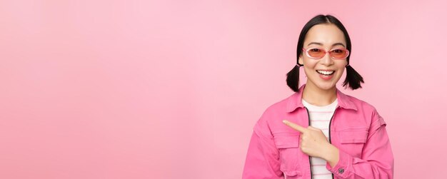 Porträt eines lächelnden asiatischen Mädchens in stilvoller Outfit-Sonnenbrille, das mit dem Finger nach links zeigt und ein Werbebanner zeigt, das über rosa Hintergrund steht